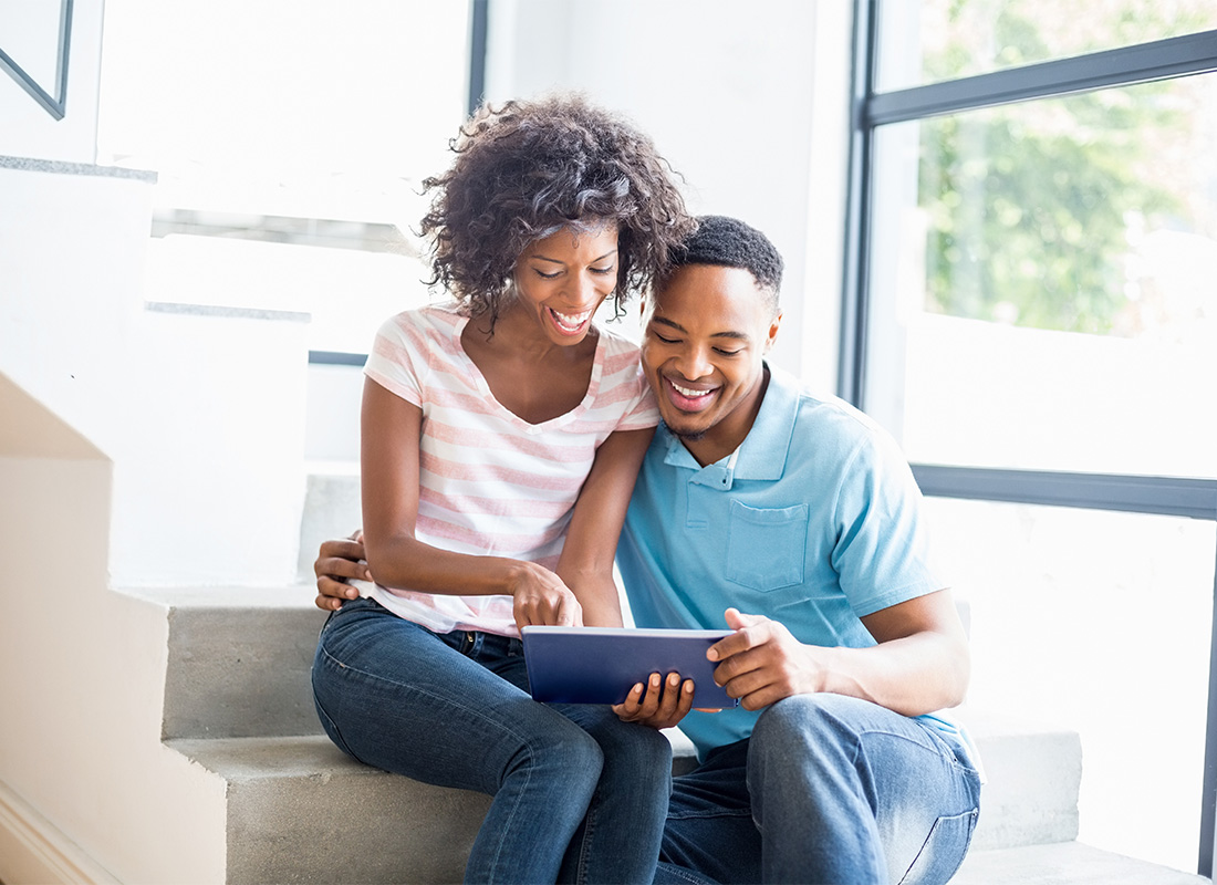 Blog - Portrait of a Cheerful Young Married African American Couple Using a Tablet While Sitting on the Stairs in Their Modern Home